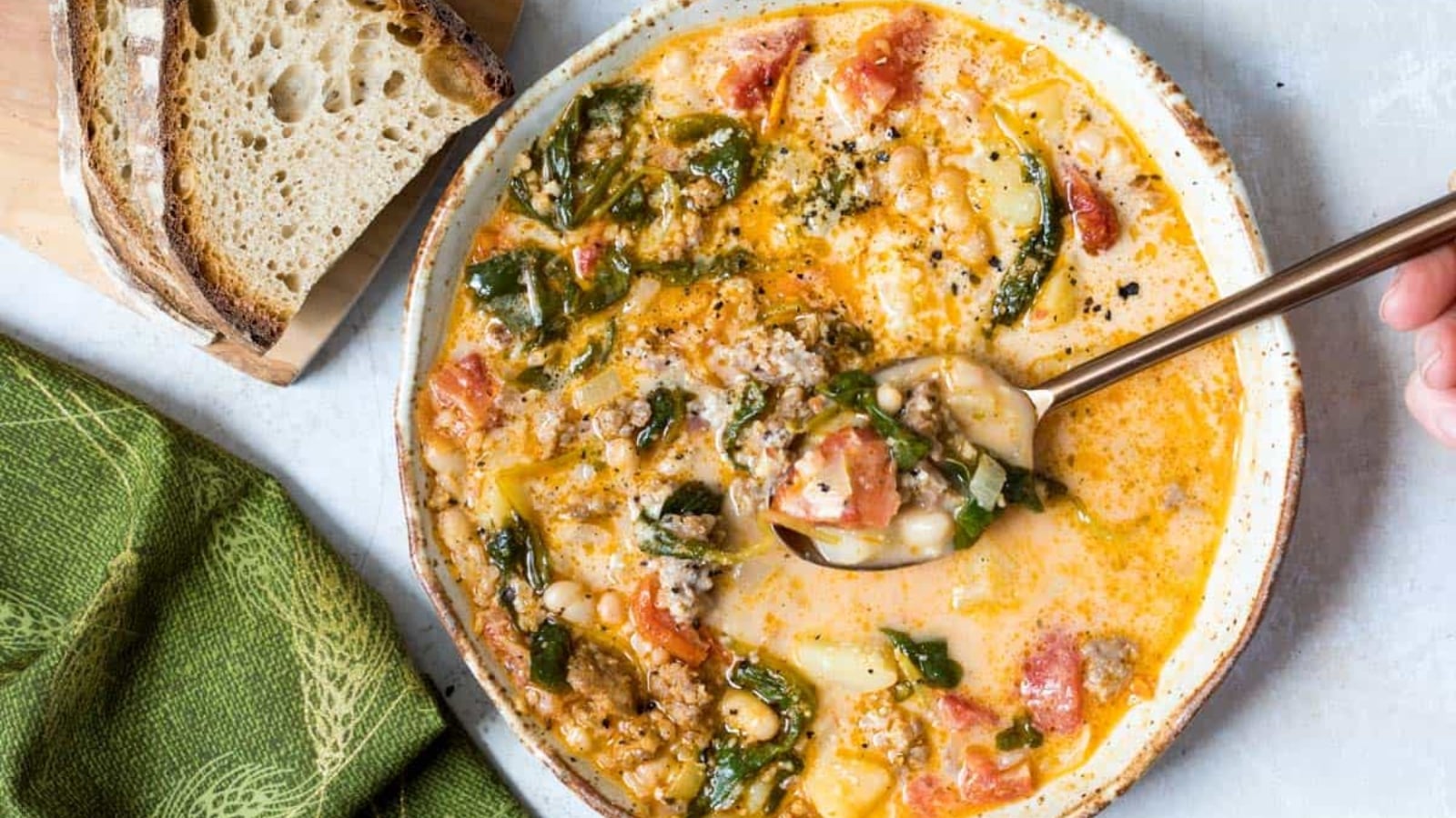 A bowl of creamy soup with sausage, white beans, spinach, and tomatoes sits next to two slices of rustic bread and a green napkin. A spoon is dipping into the soup.