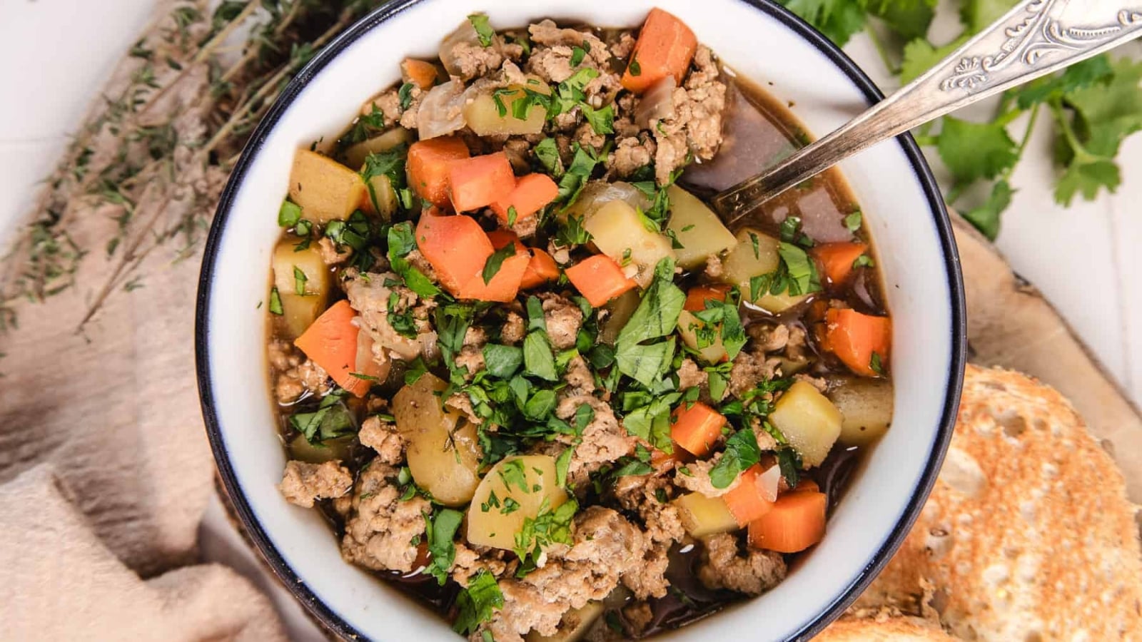 A bowl of hearty stew with ground meat, carrots, potatoes, and fresh herbs, with a spoon and bread on the side.