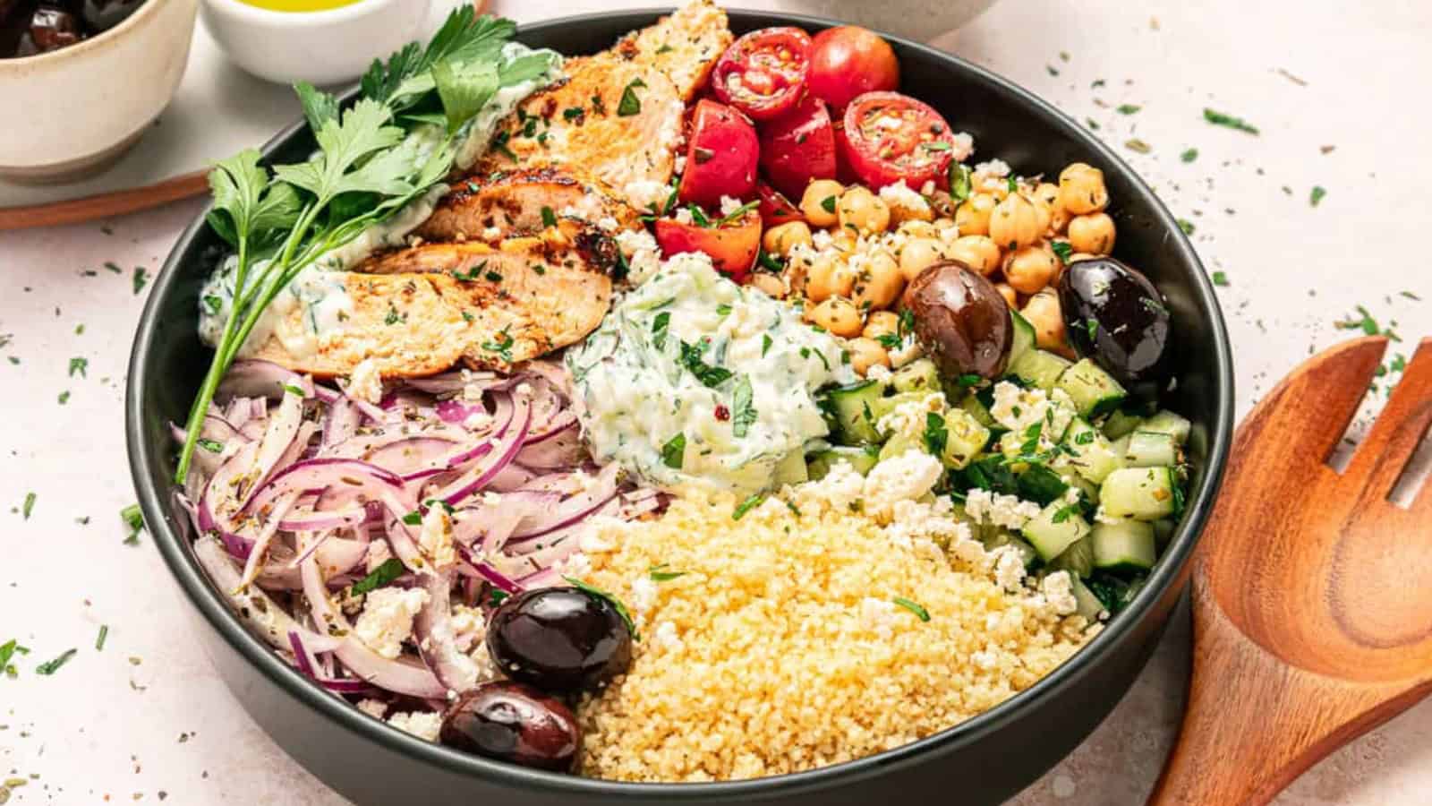 A colorful Mediterranean bowl with grilled chicken, couscous, tomatoes, cucumber, chickpeas, olives, and tzatziki.