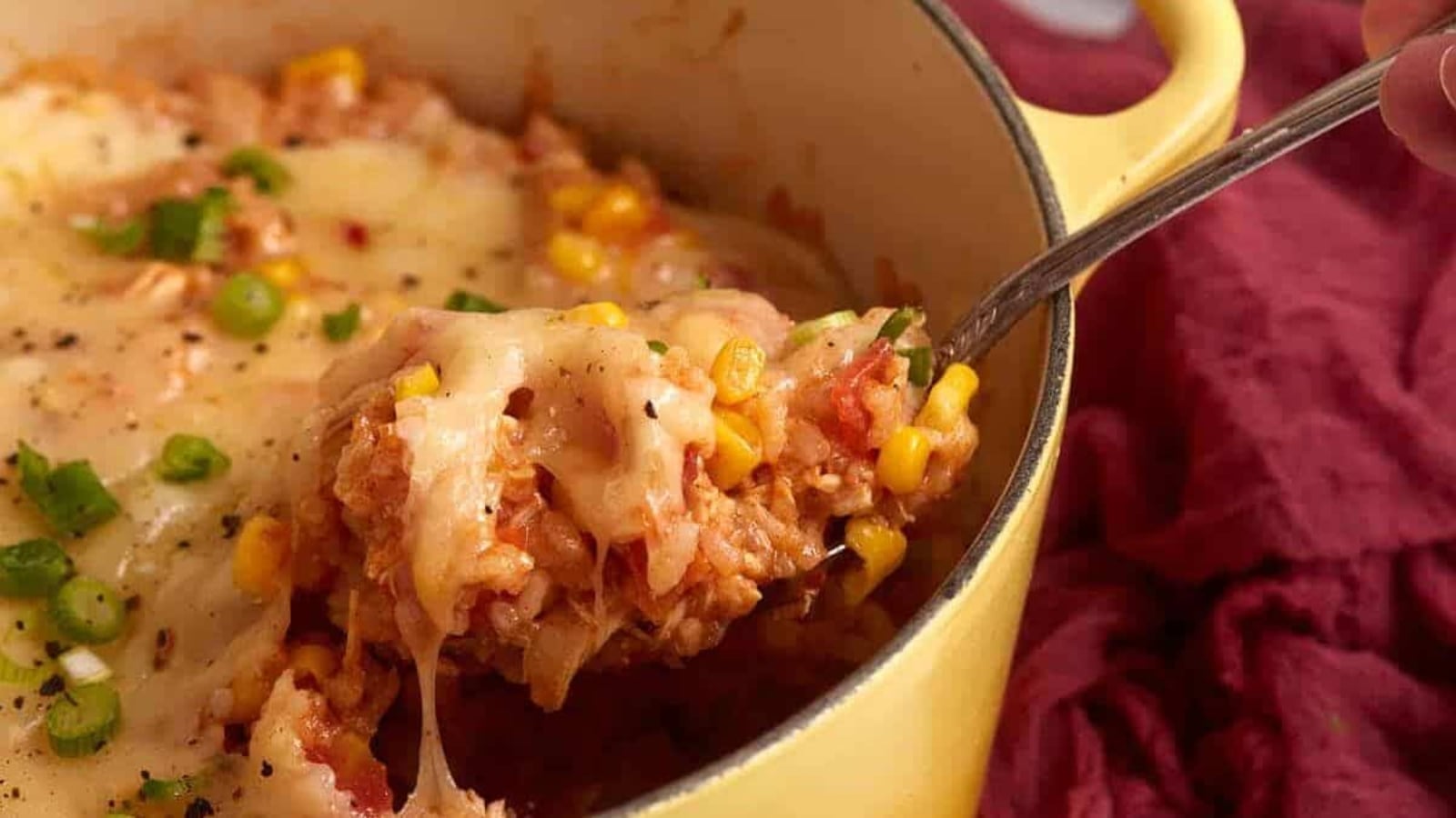 A spoon scoops cheesy rice, corn, and vegetables from a yellow pot with melted cheese on top.