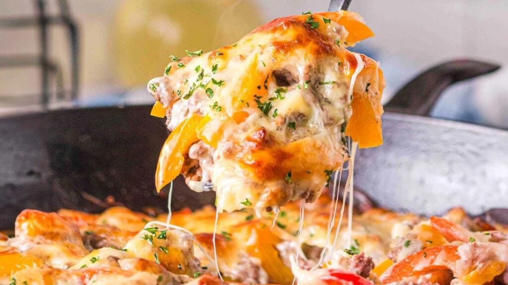 A serving of cheesy baked casserole with ground meat, bell peppers, and herbs being lifted from a skillet.