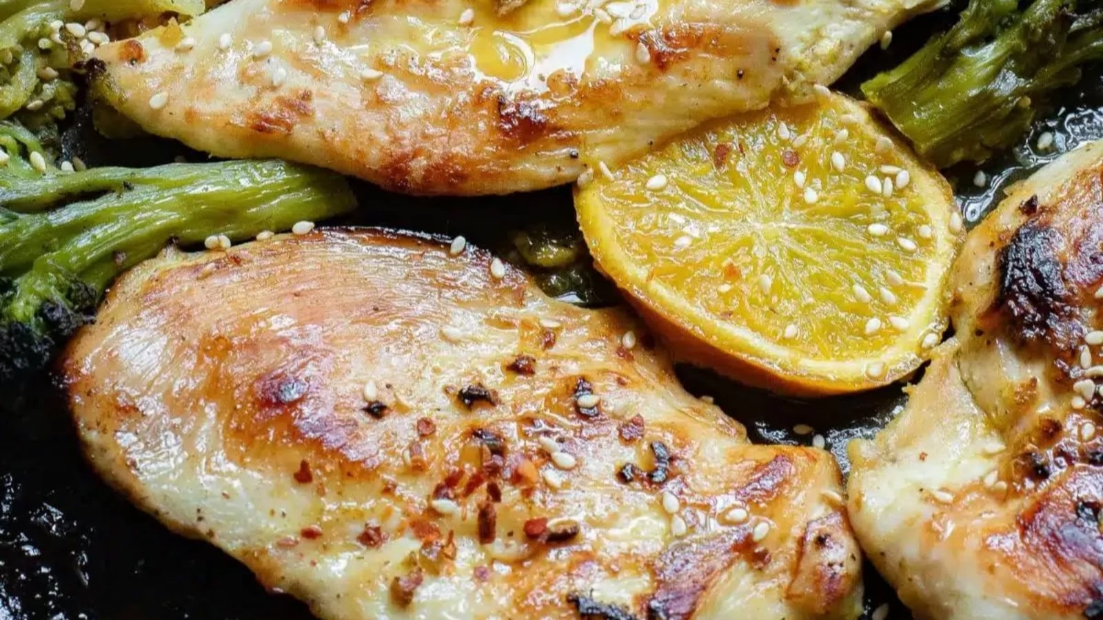 Close-up of cooked chicken breasts with herbs, red pepper flakes, and lemon slices on a bed of green vegetables.