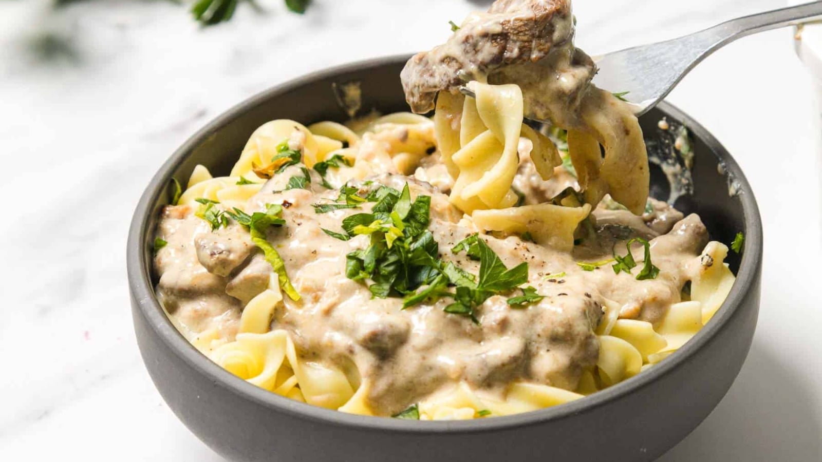 A bowl of creamy beef stroganoff with egg noodles, garnished with chopped parsley.
