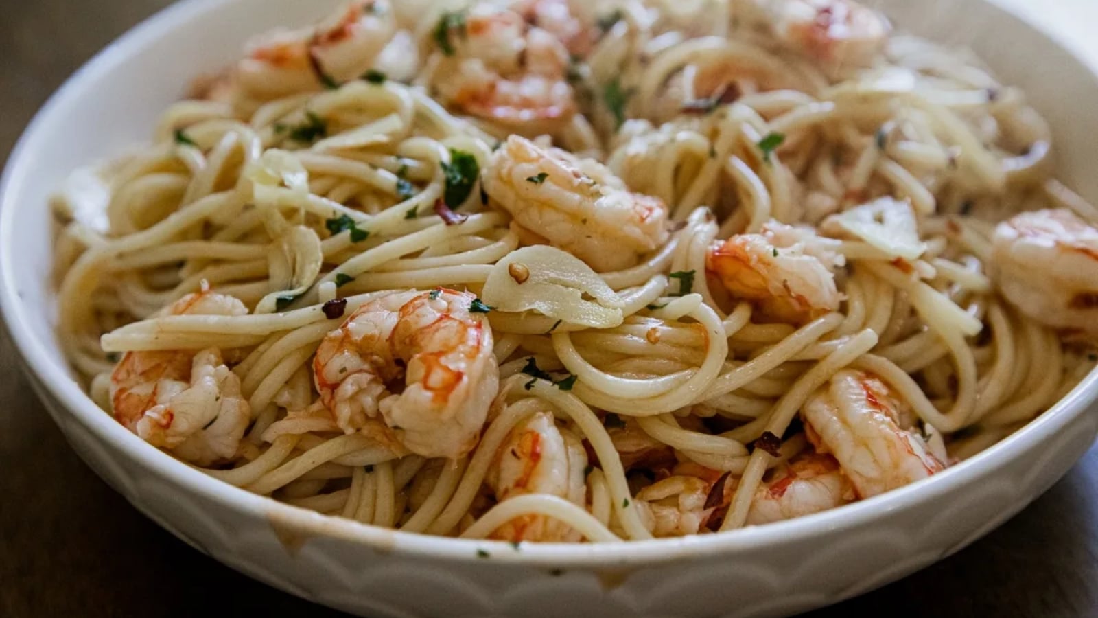 A white bowl filled with spaghetti pasta, shrimp, garlic slices, and herbs.