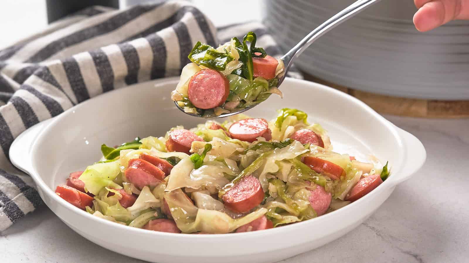 A pan filled with fried cabbage and sausage, with a wooden spoon resting inside.