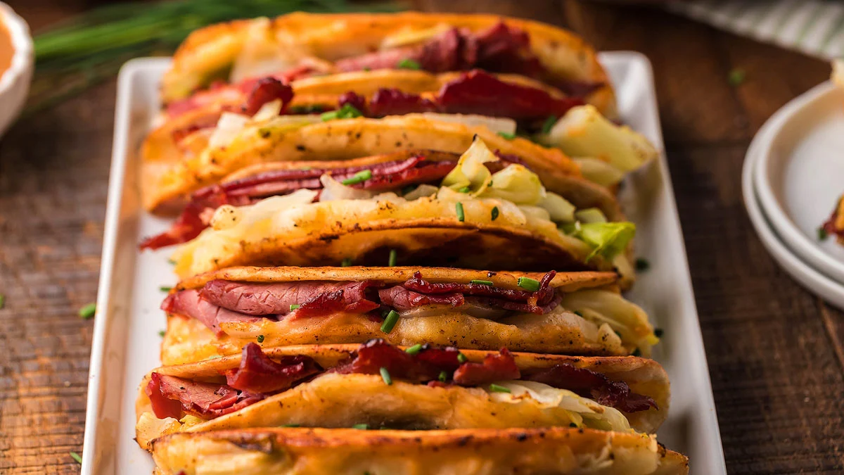 Five crispy tacos filled with shredded corned beef and cabbage are arranged on a white rectangular plate, garnished with chopped chives.