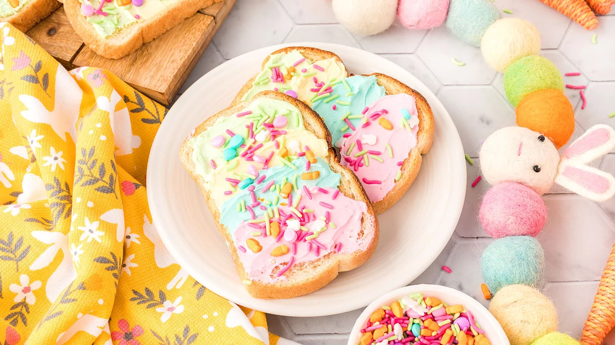 Three slices of bread topped with pastel-colored frosting and sprinkles on a white plate, next to a yellow patterned cloth and colorful felt decorations.