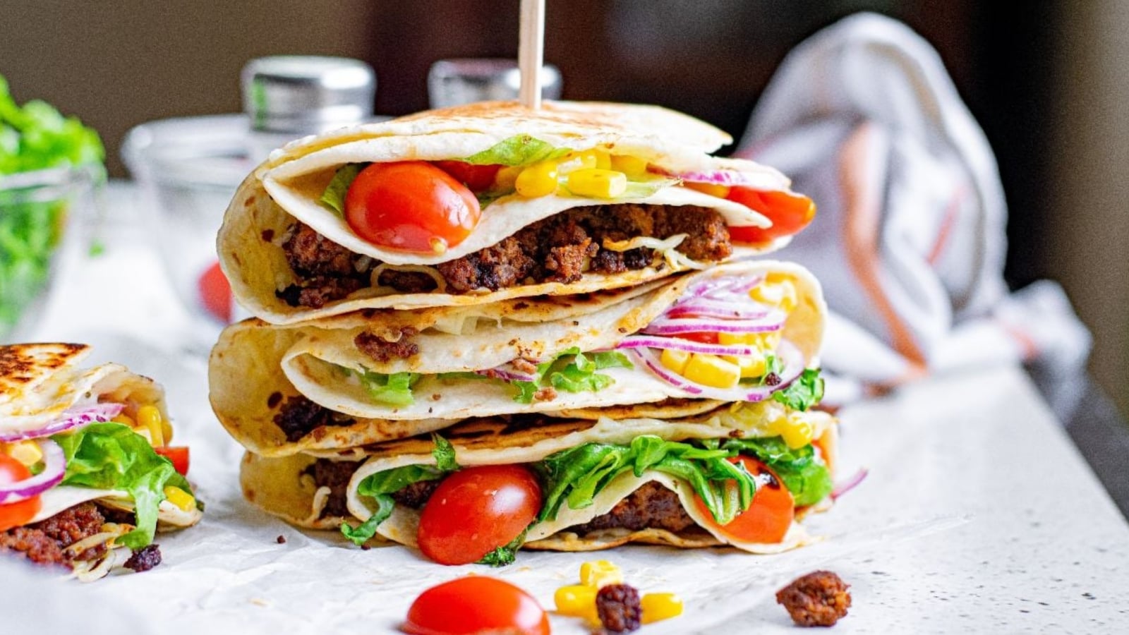 Three folded tortillas filled with ground meat, lettuce, cherry tomatoes, corn, and red onions are stacked on a countertop, with a toothpick holding the stack together.