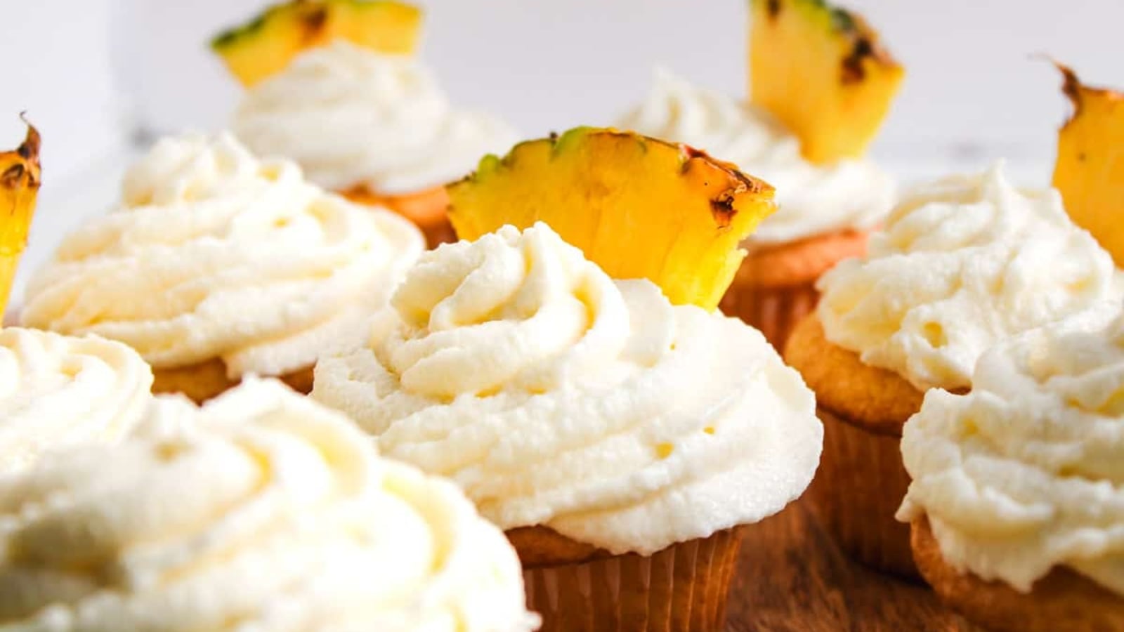 Cupcakes with swirled white frosting, some topped with pineapple slices, arranged on a wooden board.