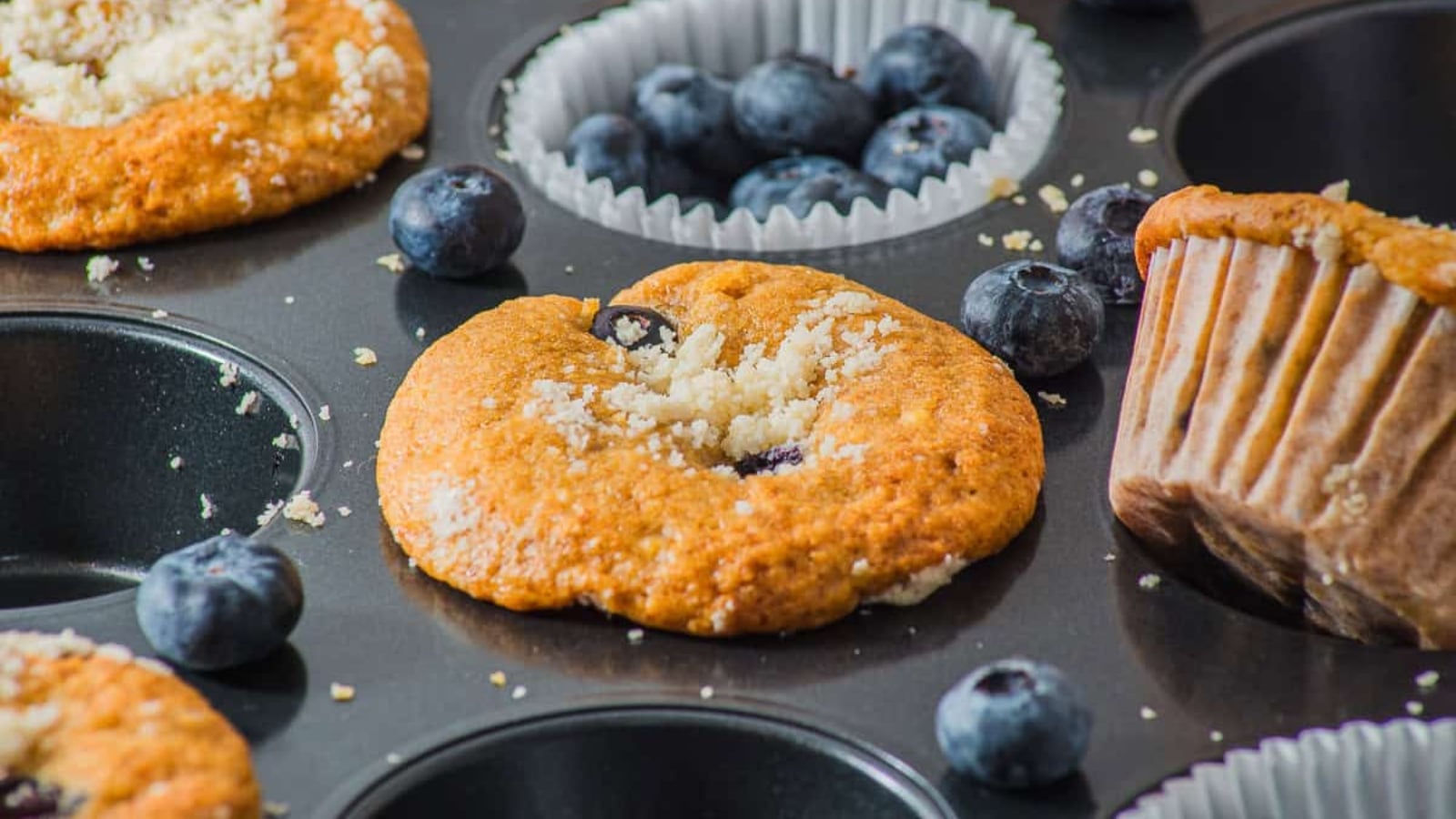 Close-up of blueberry muffins in a baking tray, with fresh blueberries and muffin crumbs scattered around.