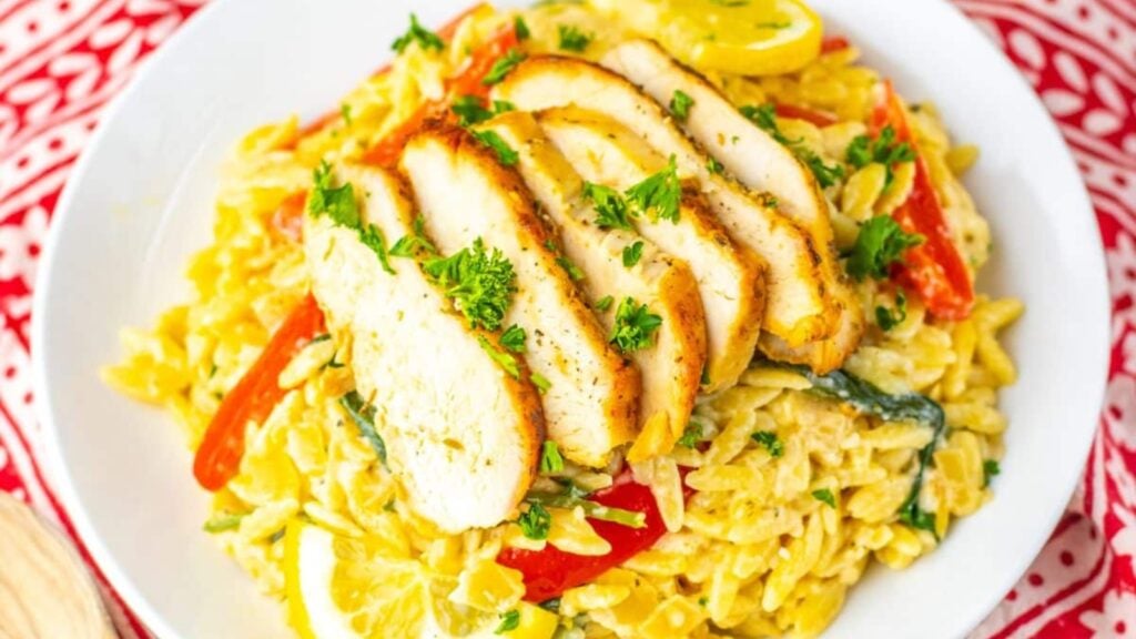A plate of creamy orzo pasta with sliced grilled chicken breast, red bell peppers, zucchini, lemon slices, and parsley garnish.