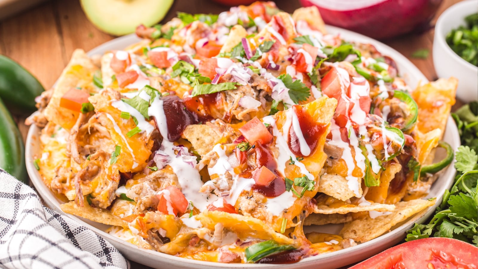 A plate of loaded nachos topped with melted cheese, diced tomatoes, chopped cilantro, barbecue sauce, and sour cream, surrounded by various fresh vegetables.