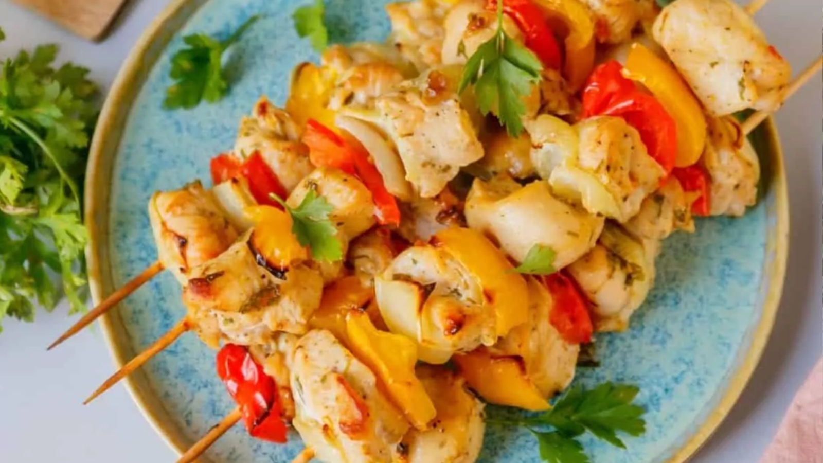 Three grilled chicken and vegetable skewers, featuring pieces of chicken, red and yellow bell peppers, and onions, are served on a blue plate with a garnish of fresh parsley.