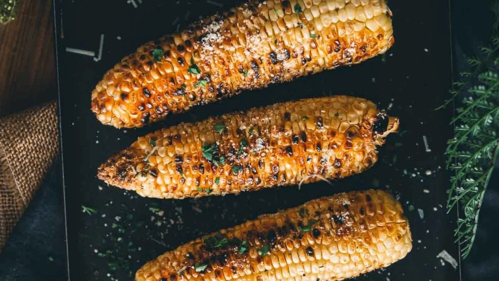 Three grilled corn cobs on a dark surface, garnished with herbs and sprinkled with grated cheese.