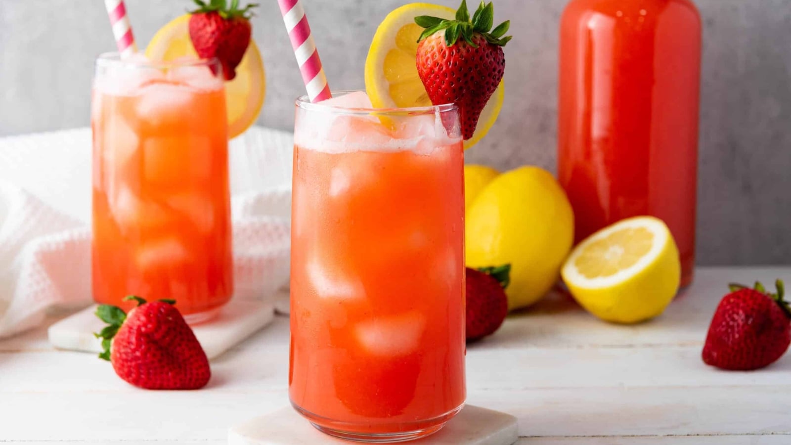 Two glasses of strawberry lemonade with ice, lemon slices, and strawberries, with striped straws on a white table.