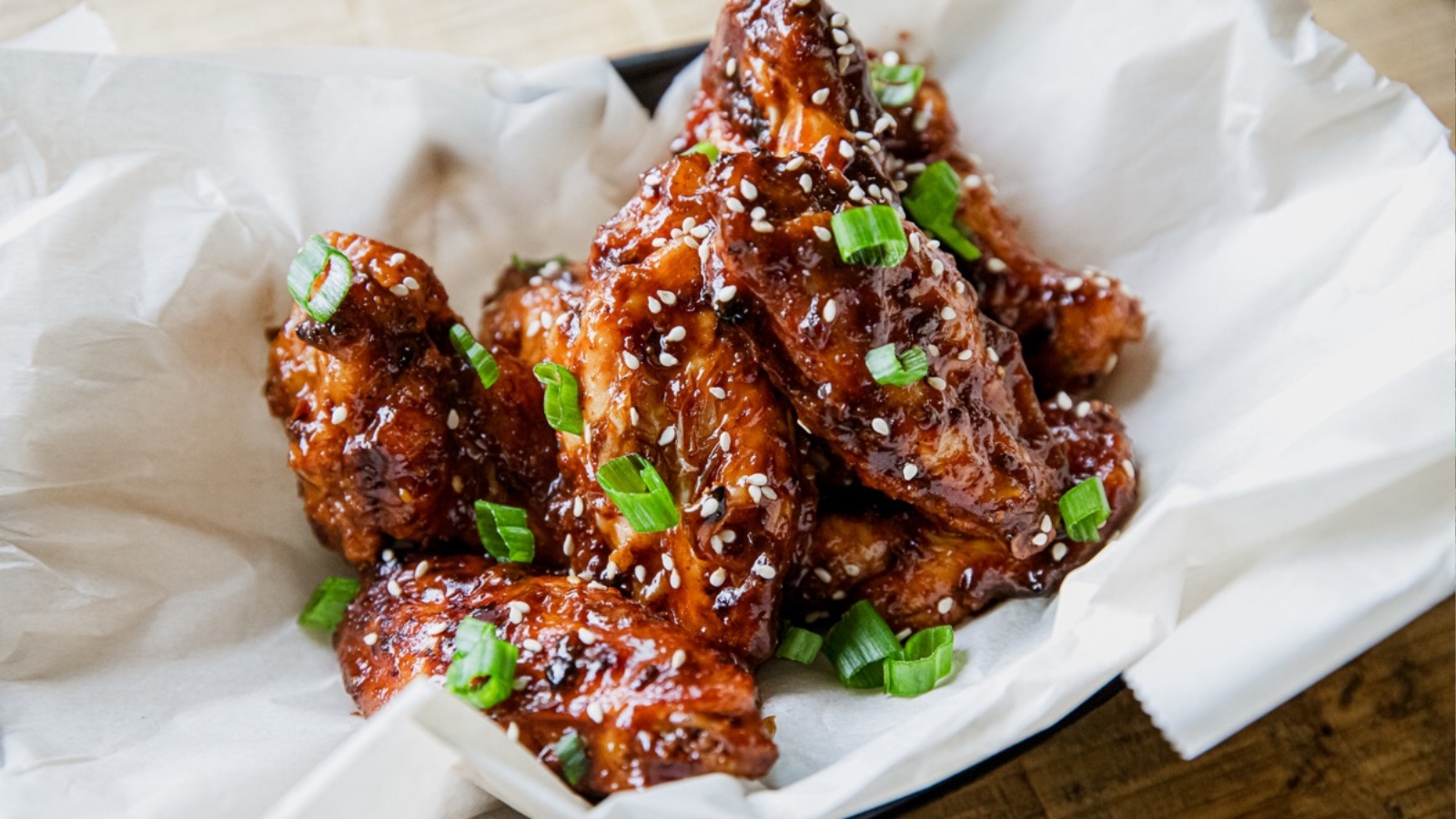 A plate of glazed chicken wings garnished with chopped green onions and sesame seeds, served on white parchment paper.