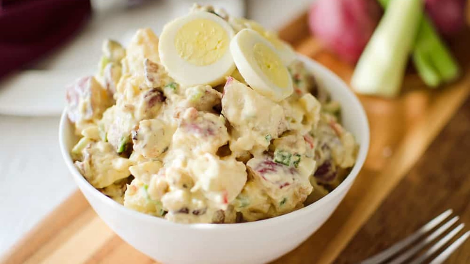 A bowl of potato salad with sliced hard-boiled eggs on top, placed on a wooden surface with vegetables in the background.