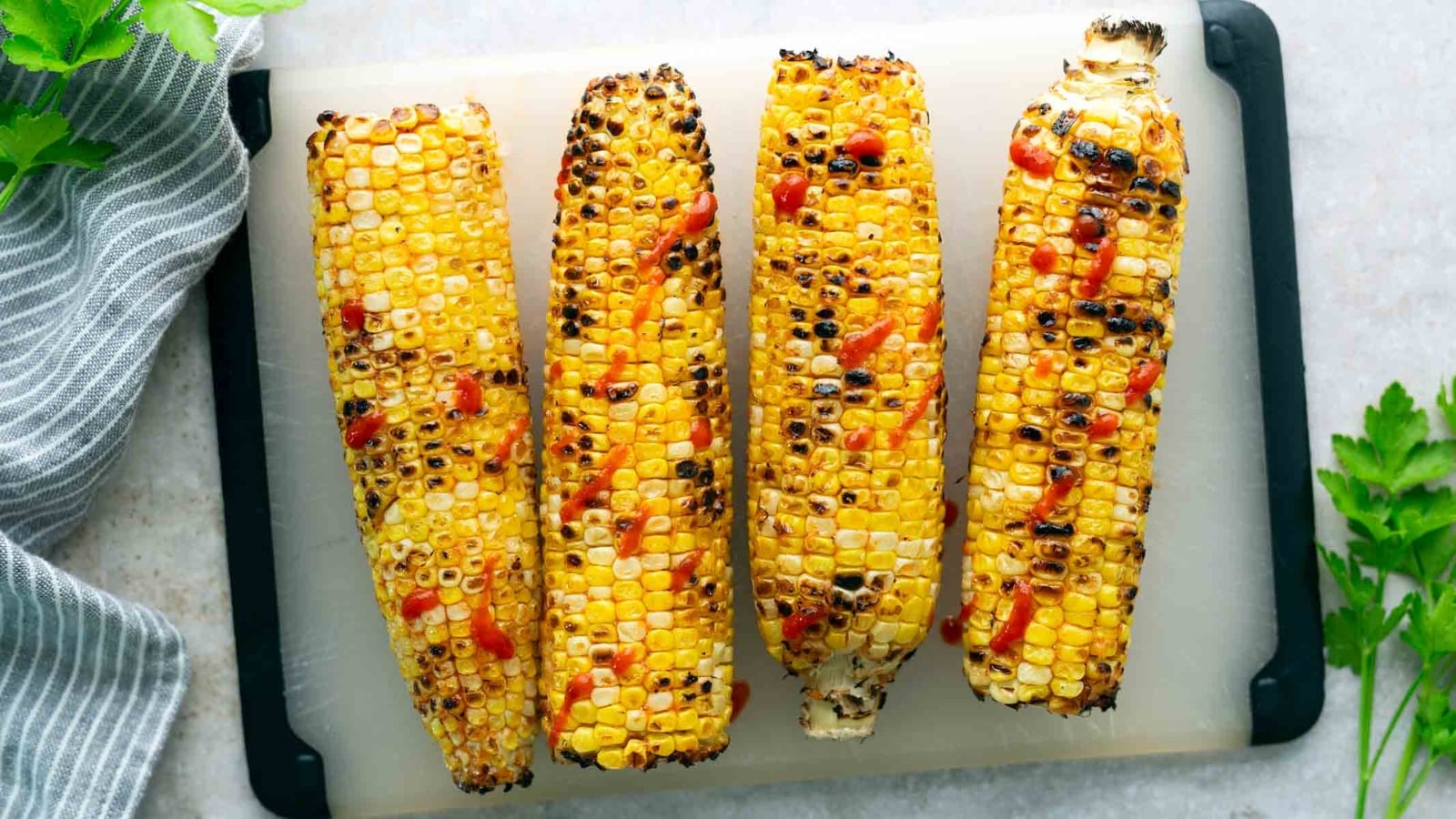 Four grilled corn cobs with char marks and a drizzle of red sauce are arranged in a row on a cutting board.