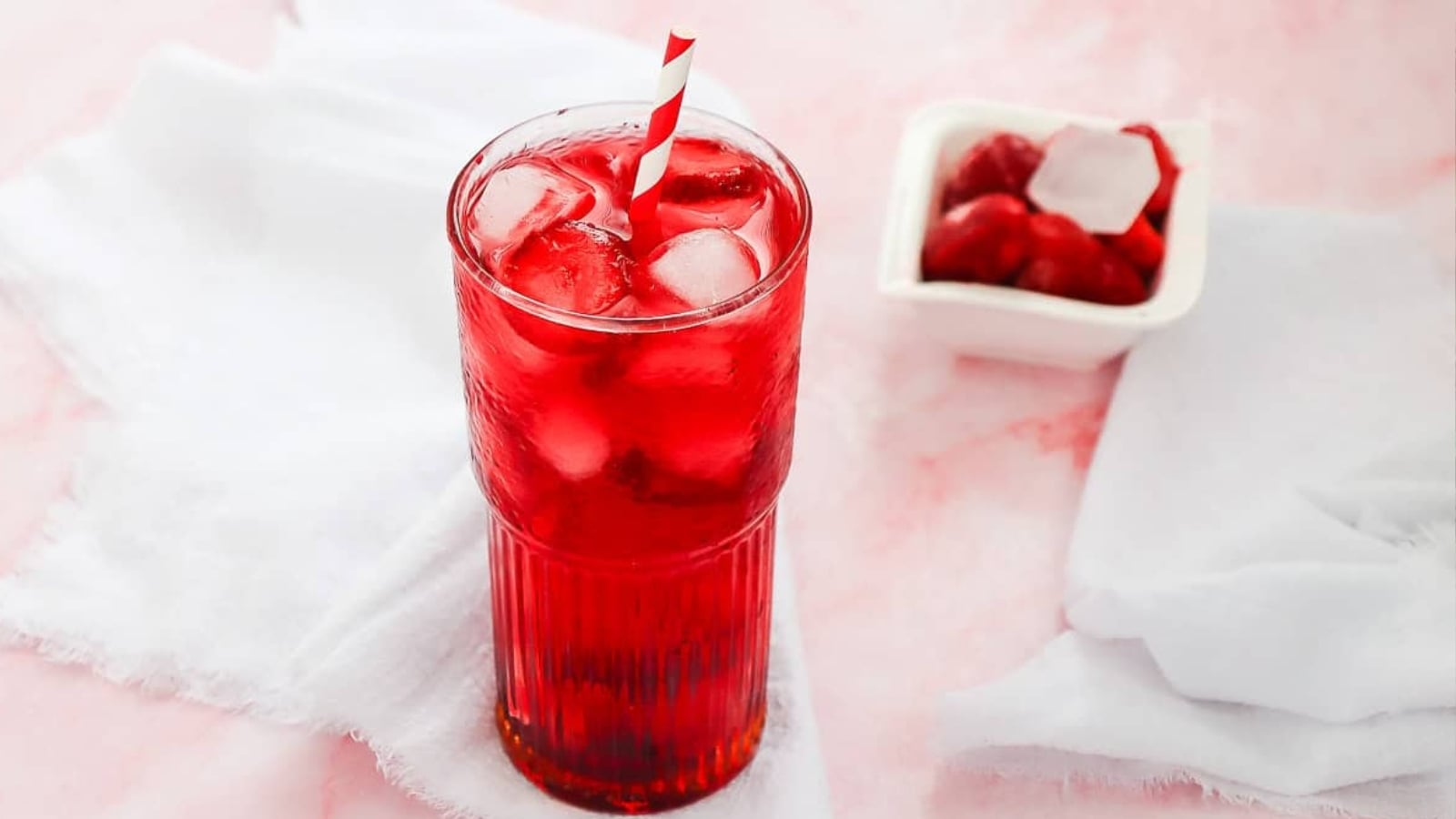 A tall glass of red iced drink with a striped straw, next to a bowl of cherries on a white cloth.