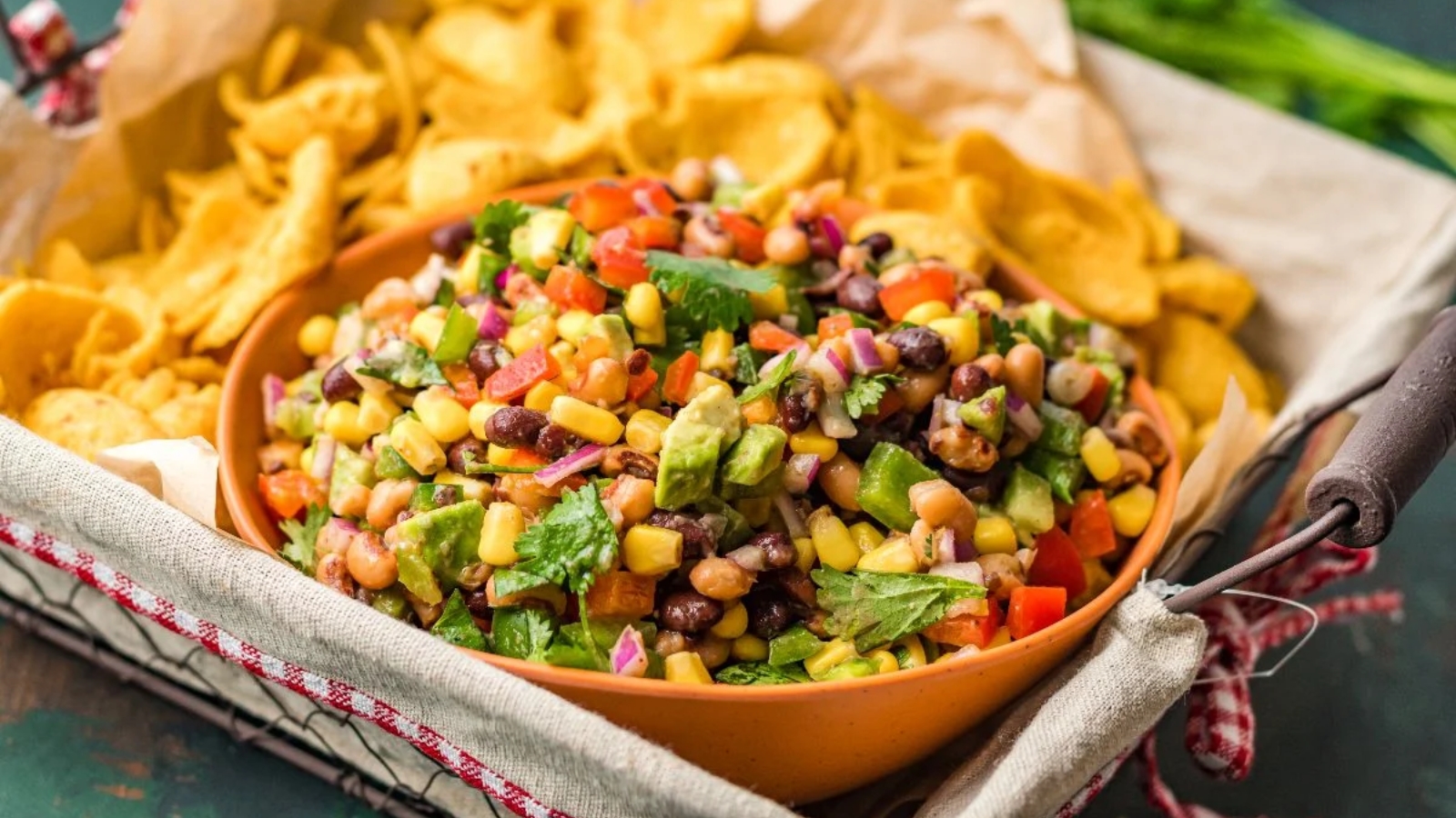 A bowl of colorful bean and corn salad with diced vegetables sits in a tray, surrounded by corn chips.