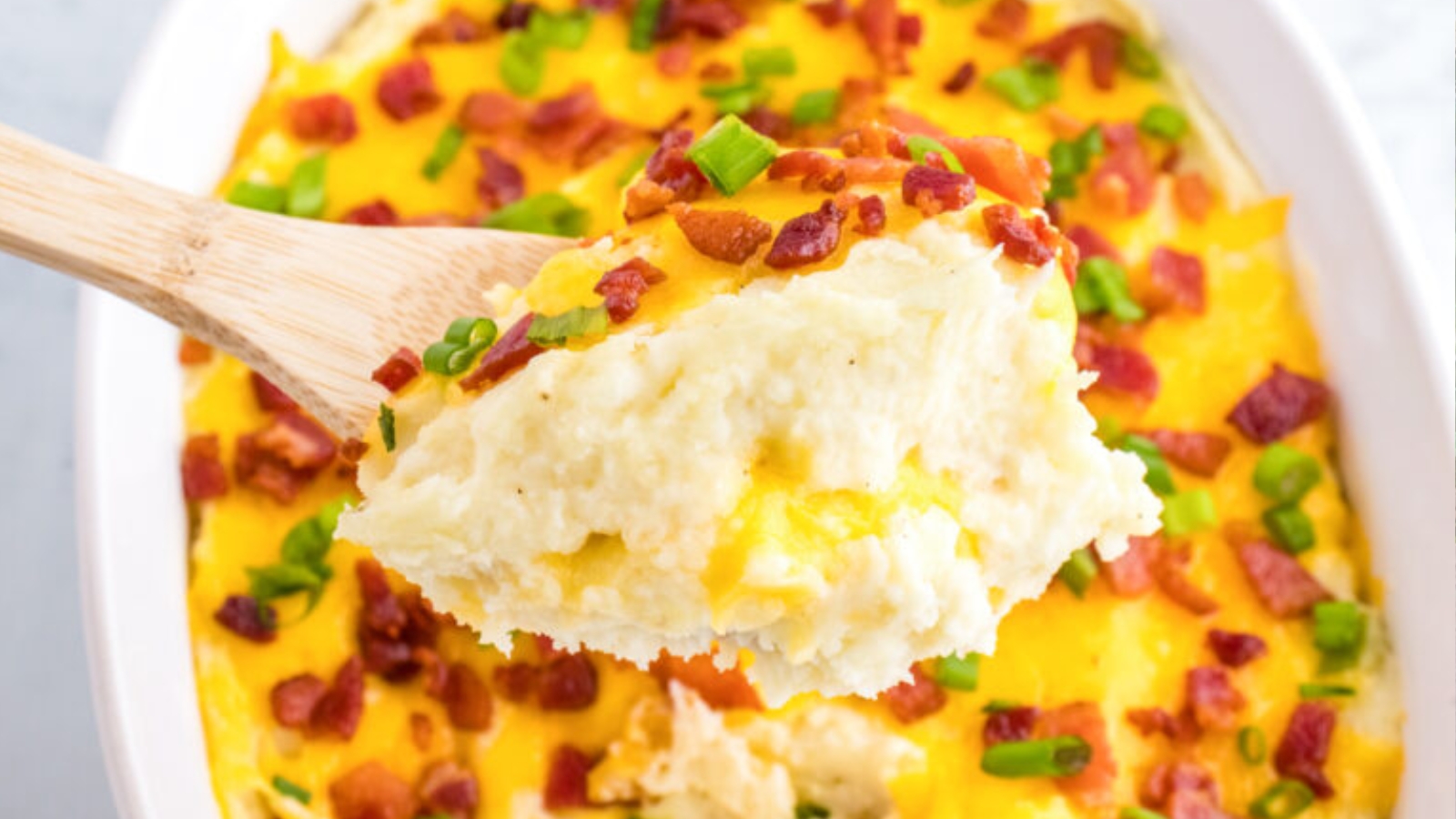 A wooden spoon holds cheesy mashed potatoes topped with bacon bits and chopped green onions.