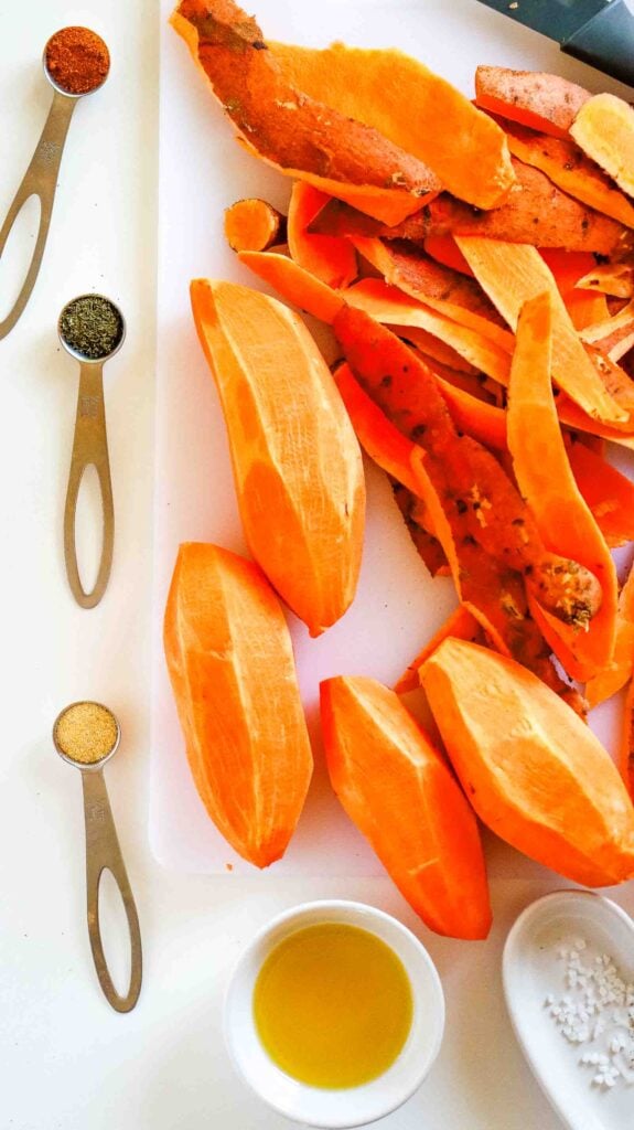 Sweet potatoes peeled and cut into even strips on a cutting board next to measured oil and seasonings.