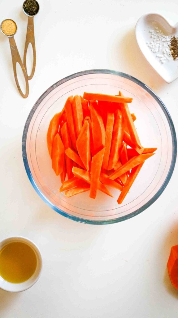 Raw sweet potato strips in a glass bowl next to measured oil, salt, and spices before tossing.