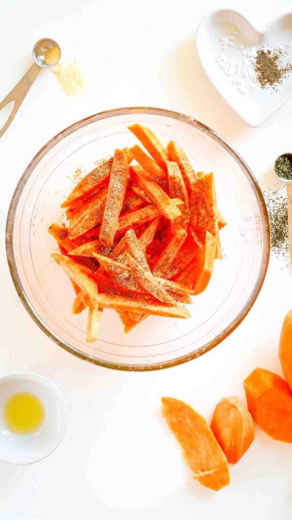 Sweet potato strips tossed and evenly coated in oil and seasoning in a glass bowl.